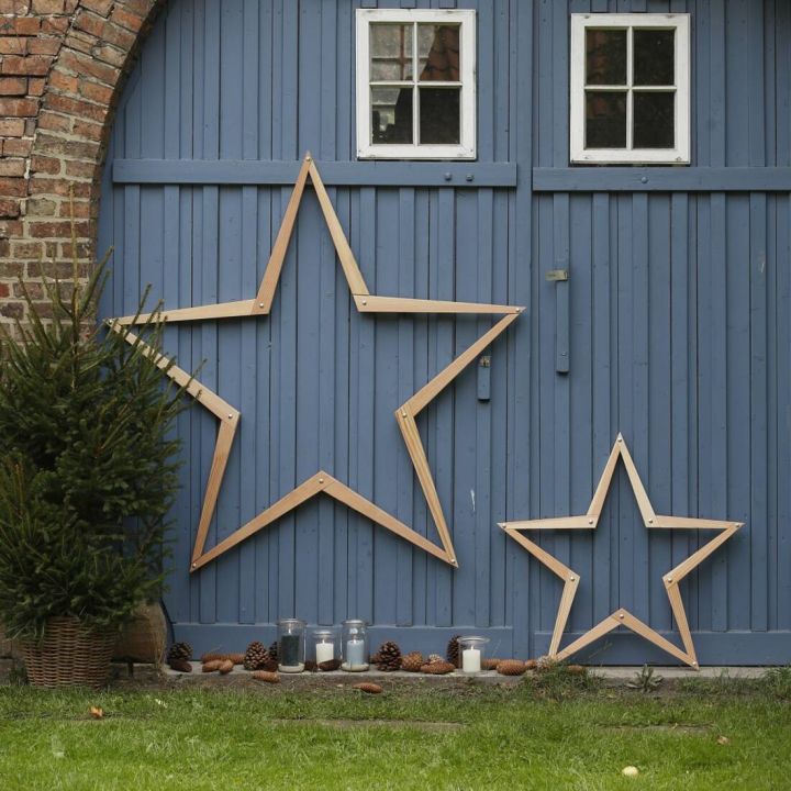 Holzsterne in zwei Größen lehnen an einer blauen Holzwand mit Fenstern. Daneben steht ein kleiner Tannenbaum im Topf. Auf dem Boden sind Kerzen in Gläsern und Tannenzapfen arrangiert.
