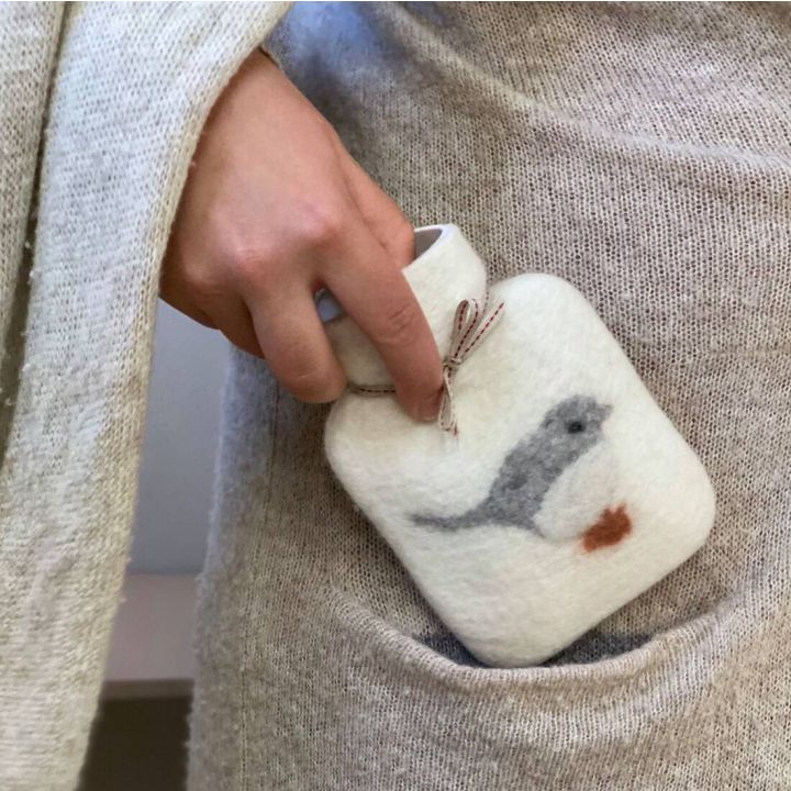 Eine Hand steckt eine kleine W&auml;rmflasche in eine Tasche. Die Flasche hat einen gestrickten &Uuml;berzug mit einem aufgestickten grauen Vogelmotiv. Die Person tr&auml;gt einen hellen Strickpullover.