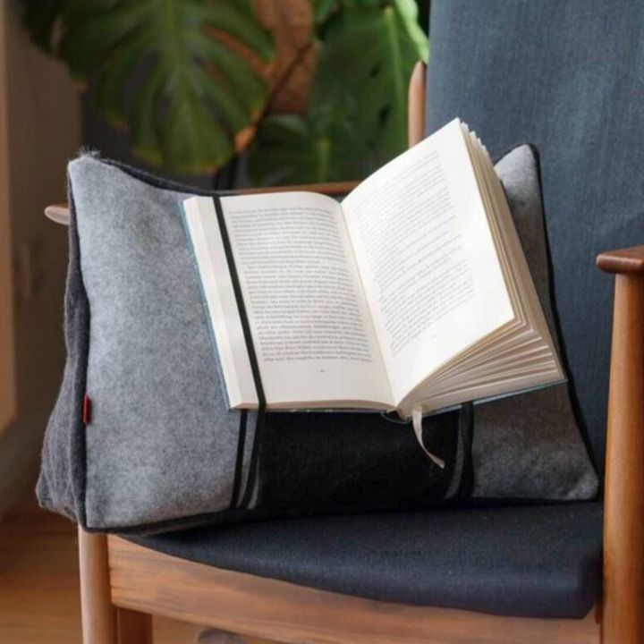 Grau-schwarzes Lesekissen mit Haltefunktion für ein geöffnetes Buch, auf einem dunkel gepolsterten Holzstuhl platziert; elastische Bänder fixieren das Buch offen.
