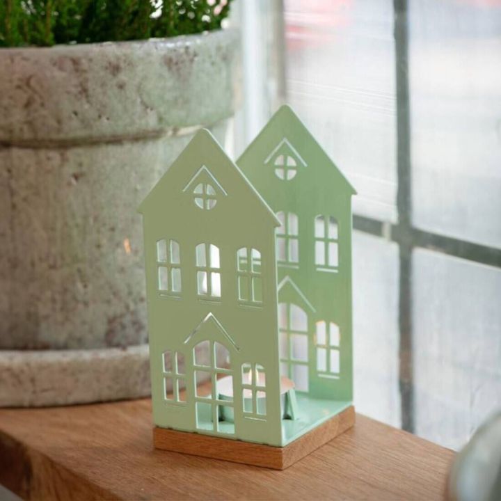 Pfefferminzgrüne Metall-Windlichtlaterne in Form eines Hauses mit mehreren Fensterausschnitten, auf einem Holzsockel; steht auf einem Holztisch neben einem großen Blumenübertopf.