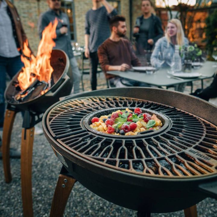 Eine Grillpfanne auf einem Outdoor-Grill, gef&uuml;llt mit Fr&uuml;chten und Minzbl&auml;ttern. Im Hintergrund sitzen mehrere Personen an einem Tisch, teils mit Getr&auml;nken, w&auml;hrend ein zweiter Grill mit Flammen zu sehen ist.