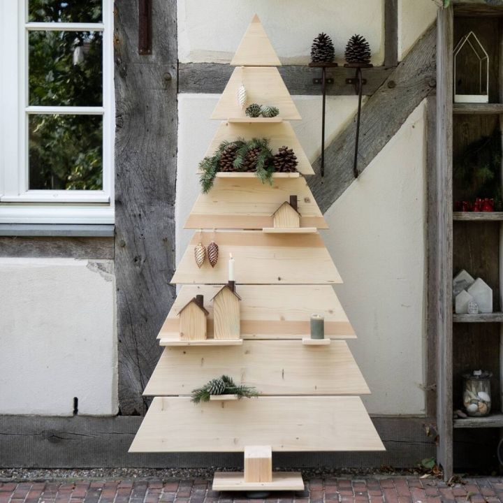 Dekorativer Weihnachtsbaum aus Holz, dreieckig gestuft, verziert mit bunten Kugeln, kleinen Figuren und Lichterkette, steht in modernem, hellen Raum mit Holzboden und halboffener Vorhangseite.