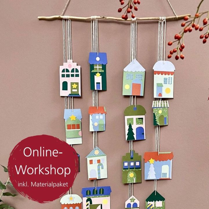 Bunte Papierhäuser auf Fäden hängend, an einem Ast befestigt. Dies ist ein kreatives Bastelprojekt, das in einem Online-Workshop angeboten wird. Rote Beschriftung „Online-Workshop inkl. Materialpaket“ links unten. Dekorative rote Beeren im Hintergrund.