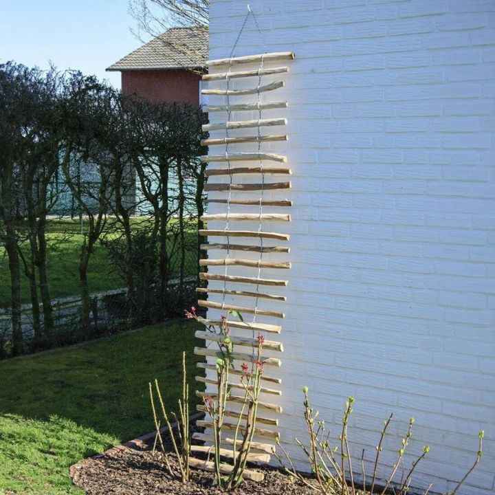 Holz-Seilleiter h&auml;ngt an wei&szlig;er Hauswand, daneben ein Garten mit Str&auml;uchern und Rasen im Hintergrund.