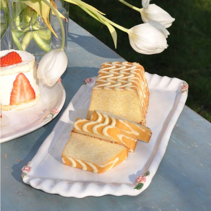 Ein rechteckiger Marmorkuchen mit Zickzackmuster, teilweise in Scheiben geschnitten, auf einem weißen Teller mit Rosenverzierungen. Links daneben eine Erdbeerrolle auf einem runden Teller. Im Hintergrund weiße Tulpen in einer Vase.