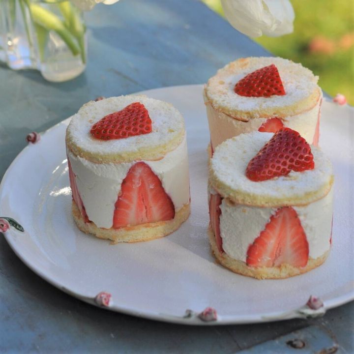 Drei runde Erdbeertörtchen auf weißem Teller. Biskuitteig, gefüllt mit Sahnecreme, mit halbierten Erdbeeren seitlich. Oben Puderzucker und Erdbeerscheiben in Herzform. Teller mit rosa Verzierungen auf blauer Oberfläche draußen.