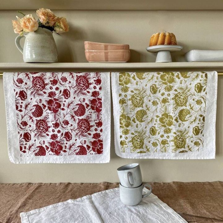 Zwei gestempelte Geschirrtücher mit Blumenmustern in Rot und Grün hängen über einer Stange, dahinter Vasen und Backform auf Regal, daneben Tassen auf Tischdecke.