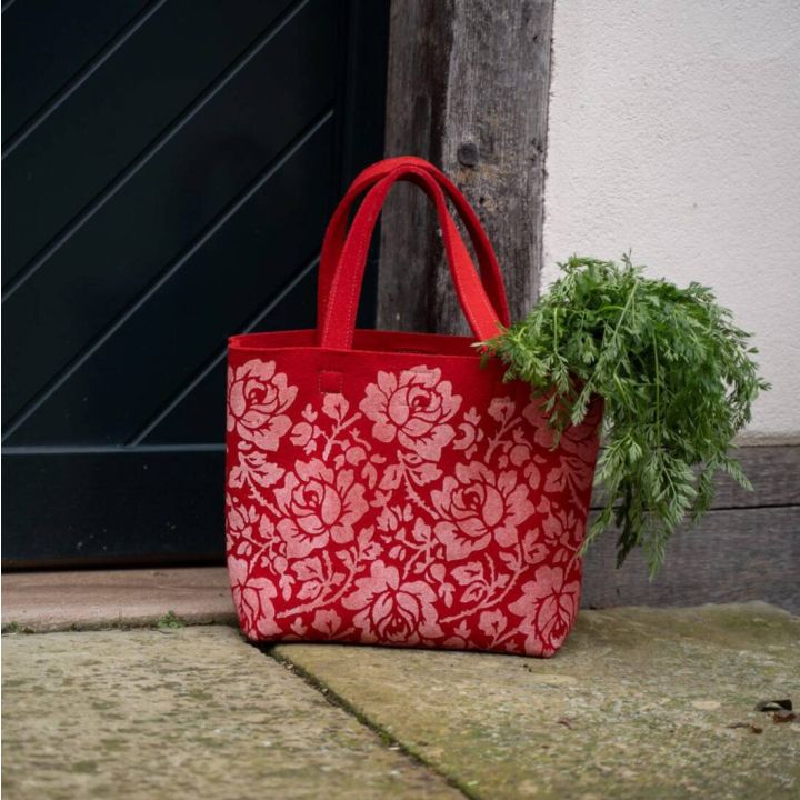 Eine rote Stofftasche mit floralen Mustern, gefüllt mit Karottengrün, steht auf einem Steinboden vor einer dunklen Tür und einer Holzwand.