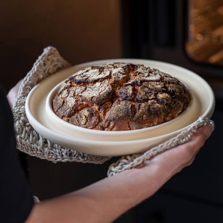 Eine Person hält ein rundes, gebackenes Brot auf einem weißen Backstein. Das Brot hat eine knusprige, rissige Kruste und wird mit Topflappen aus Strickstoff gehalten. Im Hintergrund ist ein geöffneter Backofen erkennbar.
