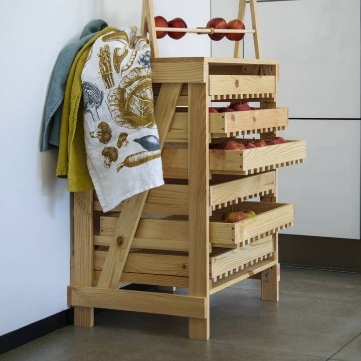 Holzregal mit vier offenen Schubladen, gef&uuml;llt mit roten &Auml;pfeln. Auf der linken Seite h&auml;ngt ein bunt bedrucktes Geschirrtuch. Das Regal steht auf einem Fliesenboden neben einer wei&szlig;en Wand.