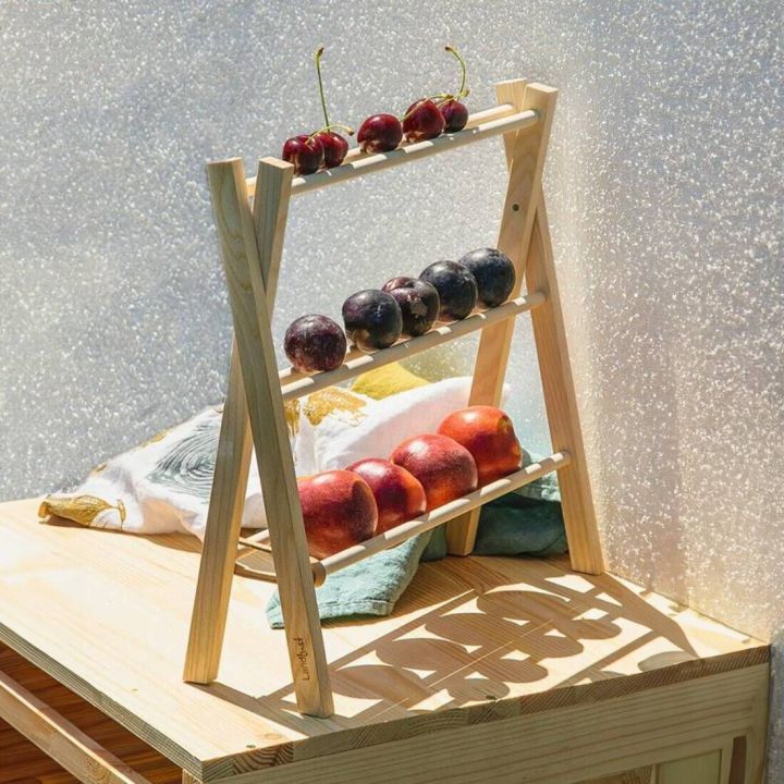 Holzregal in A-Form mit drei Ebenen, auf denen Kirschen, Pflaumen und Nektarinen liegen; steht auf einem Holztisch, dekoriert mit Kissen und einer Stoffserviette in Sonnenlicht.