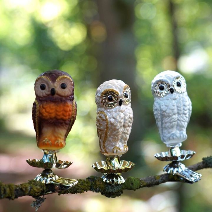 Drei dekorative Eulenfiguren in verschiedenen Farben stehen auf einer Astkerze, jede montiert auf einem metallenen Clip. Der Hintergrund ist ein verschwommener Wald.