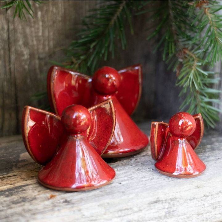 Drei rote, glasierte Keramikengel in unterschiedlichen Größen stehen auf einem hölzernen Untergrund, umrahmt von grünen Tannenzweigen.