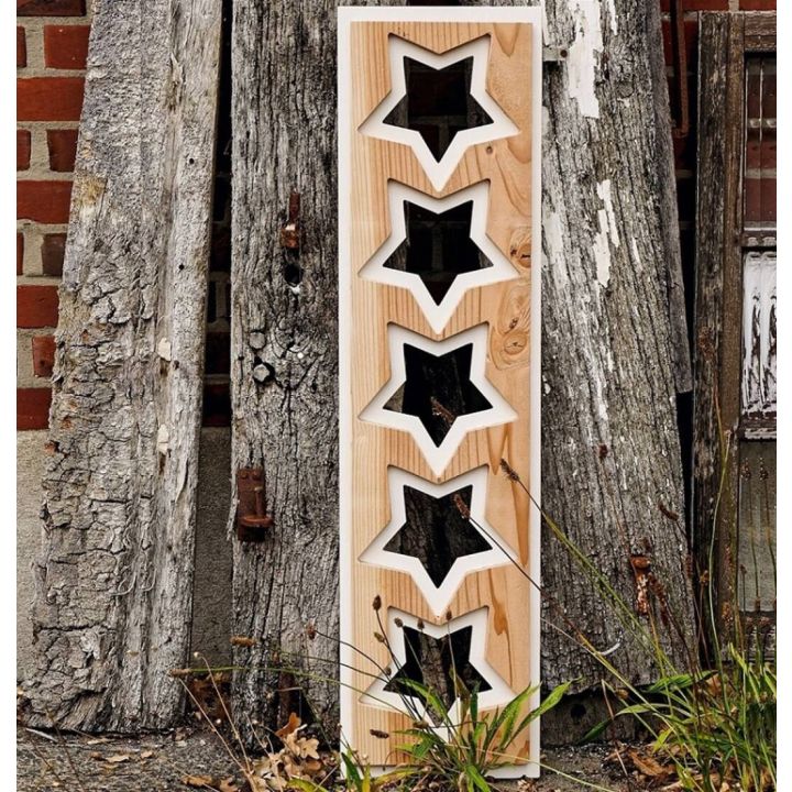 Ein rechteckiges Holzbrett mit f&uuml;nf sternf&ouml;rmigen Ausschnitten steht aufrecht drau&szlig;en, gest&uuml;tzt von verwittertem Holz vor einer Ziegelwand.