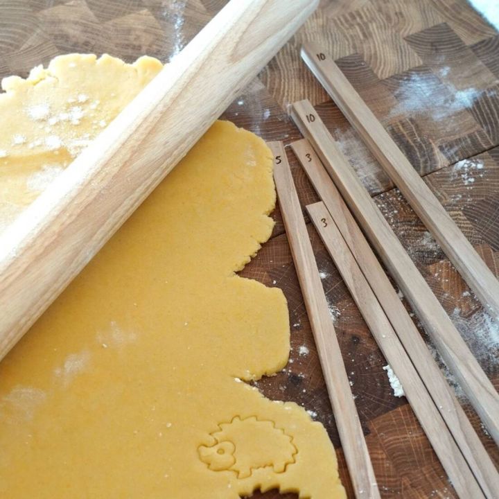 Ausgerollter gelber Teig auf einem hölzernen Schneidebrett, teilweise mit einem Nudelholz bearbeitet. Daneben liegen mehrere Holzleisten mit eingravierten Zahlen (3, 5, 10), die als Abstandshalter zur gleichmäßigen Teigstärke dienen. Im Teig ist eine Auss