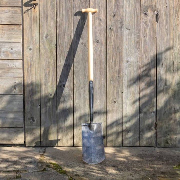 Garten-Spaten mit T-Griff aus hellem Holz und schwarzem Metallblatt, aufrecht an eine Holzwand gelehnt, vollständig sichtbar, auf Steinboden stehend, fotografiert bei Tageslicht.