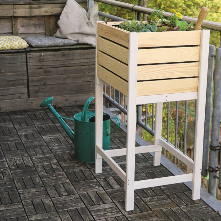Hochbeet aus Holz auf einem Balkon mit Gittern, daneben eine grüne Gießkanne auf Holzbodenfliesen. Im Hochbeet sind Pflanzen sichtbar, im Hintergrund liegt eine Sitzbank mit Kissen.