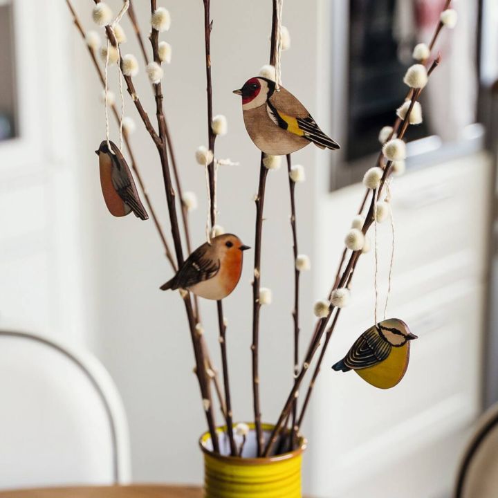 Zweige in gelber Vase mit weißen Knospen und vier hängenden Vogelholzdekorationen in verschiedenen Farben und Designs.