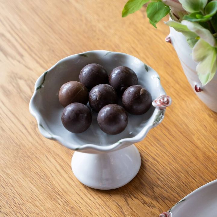 Weiße Porzellanschale auf Sockel mit sieben dunklen Pralinen, arrangiert auf einem Holztisch. Daneben sind Teile einer dekorierten Vase mit grünen und rosafarbenen Blumen sichtbar.