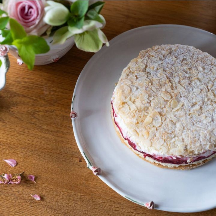 Eine runde Torte mit Mandelbl&auml;ttchen auf der Oberfl&auml;che und einer roten Fruchtschicht in der Mitte auf einem wei&szlig;en Teller, umgeben von Blumen im Hintergrund auf einem Holztisch.