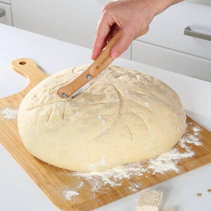 Hand ritzt mit einem speziellen Teigmesser einen runden Brotteig auf bemehltem Holzbrett ein; Messer mit Holzgriff und gebogener Klinge, Teig mit dekorativen Einschnitten vorbereitet.