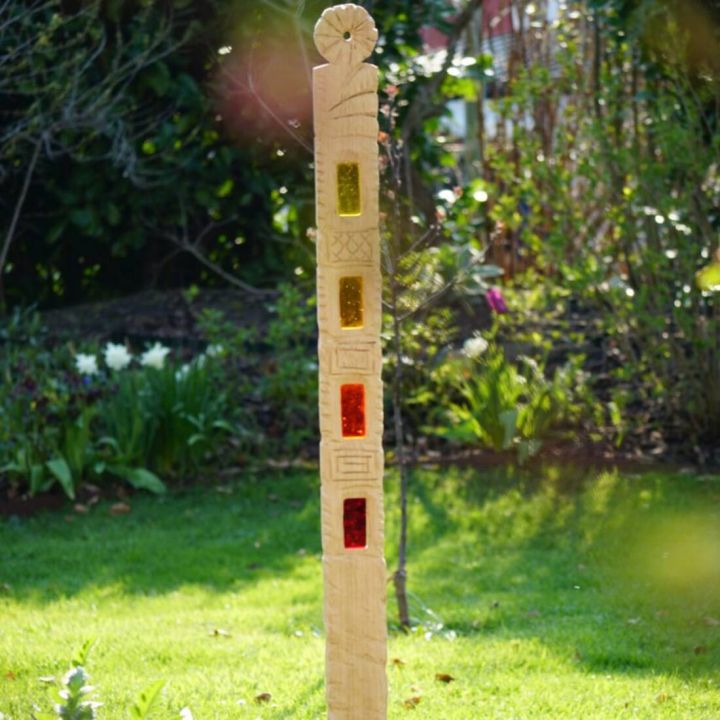 Aufrecht stehender hellholzfarbener Sonnenf&auml;nger, ca. 1 m hoch, oben runde Rosette, darunter vier vertikale rechteckige Glasfenster in Gelb-Orange-Rot, umlaufend ornamental geschnitzt; fotografiert freistehend auf Rasen mit unscharfem Gartenhintergrund.