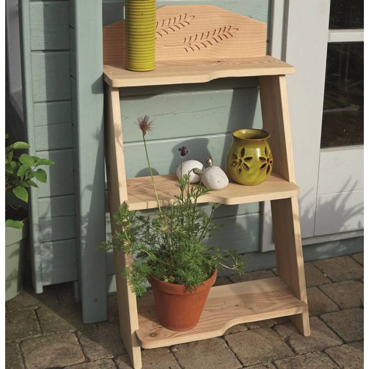 Holzregal mit drei Regalböden, dekoriert mit einem gelben Kerzenhalter, weißen Dekoeiern und einem Blumentopf mit grüner Pflanze. Im Hintergrund eine hellblaue Wand und eine weiße Tür. Standort auf Pflastersteinen.