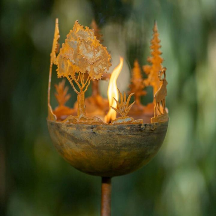 Metallische Feuerschale mit ausgestanzten Baum- und Tiermotiven, entzündet, auf Stab montiert, vor unscharfem Hintergrund.