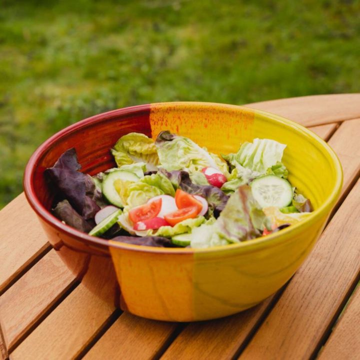 Bunte Keramikschüssel mit rot-gelbem Farbverlauf, gefüllt mit gemischtem Salat aus grünem und rotem Blattsalat, Gurkenscheiben, Tomaten- und Paprikastücken, steht auf einem Holztisch im Freien vor grünem Rasen.