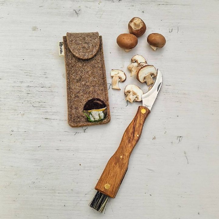 Pilzmesser mit Holzgriff und Reinigungsbürste, daneben geöffnete Filztasche; aufgeschnittene braune Champignons auf weißem Hintergrund.