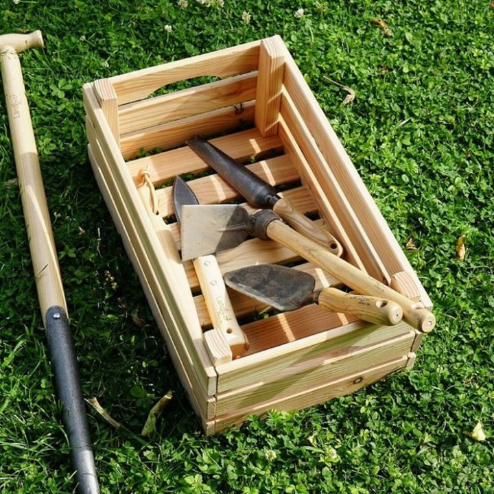 Holzkiste mit Gartenger&auml;ten (Hacke, Schaufel, Spaten) auf Rasen. Links ist ein weiteres langes Gartenger&auml;t. Die Kiste hat Seitengriffe und ist aus hellem Holz, die Werkzeuge haben Holzgriffe und metallene K&ouml;pfe.
