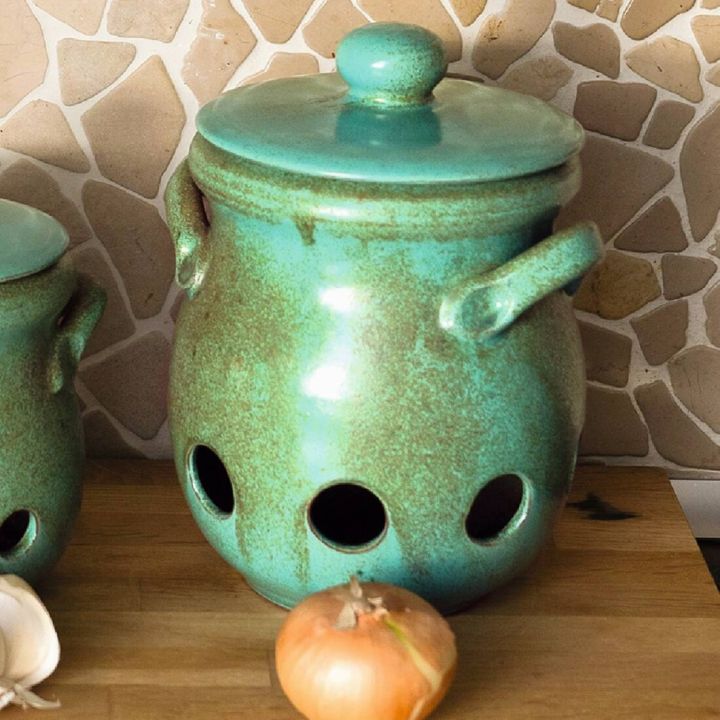 Grüner, keramischer Topf mit Deckel und Griffen, steht auf Holztisch. Vorderseite hat große, runde Löcher. Navi stabile Steinmosaikhintergrund. Eine Zwiebel liegt davor.
