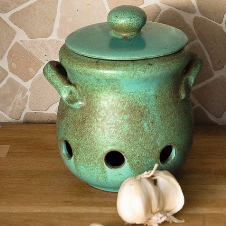 Gr&uuml;ner Keramik-Topf mit Deckel und seitlichen Griffen, hat Bel&uuml;ftungsl&ouml;cher und steht auf einer Holzoberfl&auml;che vor einer Steinwand. Zwei Knoblauchknollen liegen davor.
