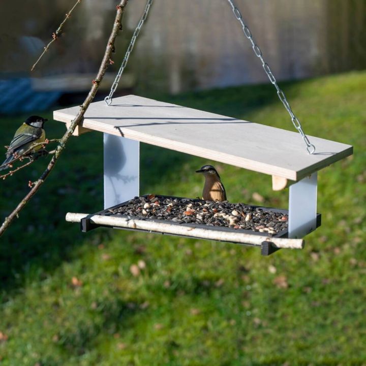 H&auml;ngendes Vogelhaus aus Holz mit Metallketten, gef&uuml;llt mit Vogelfutter, zwei kleine V&ouml;gel sitzen am Futterhaus. Im Hintergrund Rasen und B&auml;ume.