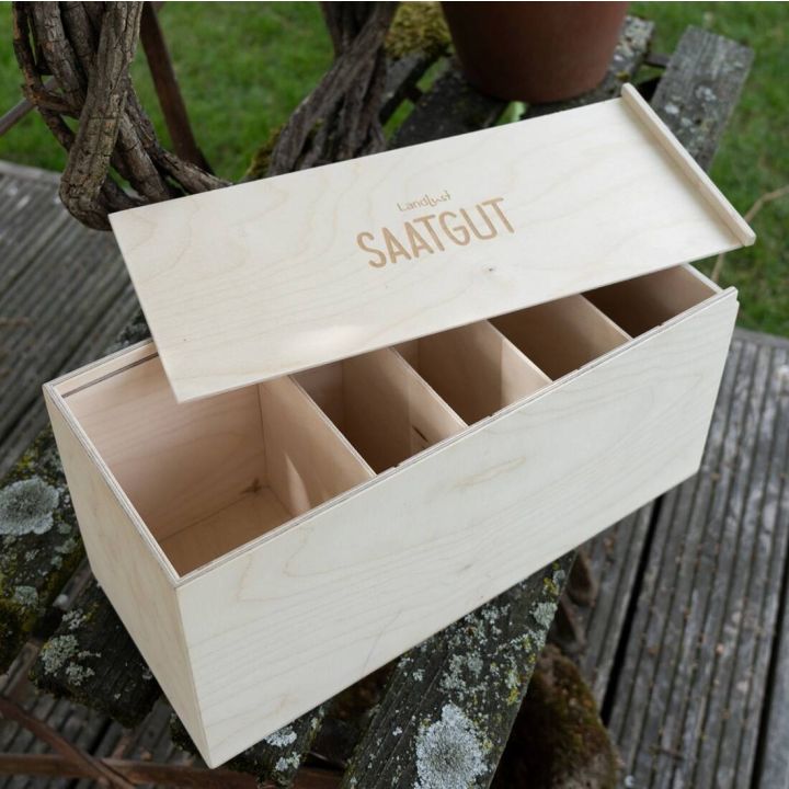 Holz-Saatgutkiste mit f&uuml;nf F&auml;chern und aufgesetztem Deckel, auf dem "Landlust SAATGUT" steht; steht auf Holztisch im Freien.