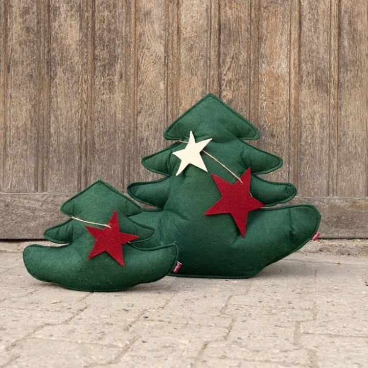 Zwei grüne Kissen in Tannenbaumform vor Holzwand. Das größere Kissen hat einen weißen und einen roten Stern, das kleinere einen roten Stern. Kissen stehen auf einem Boden.