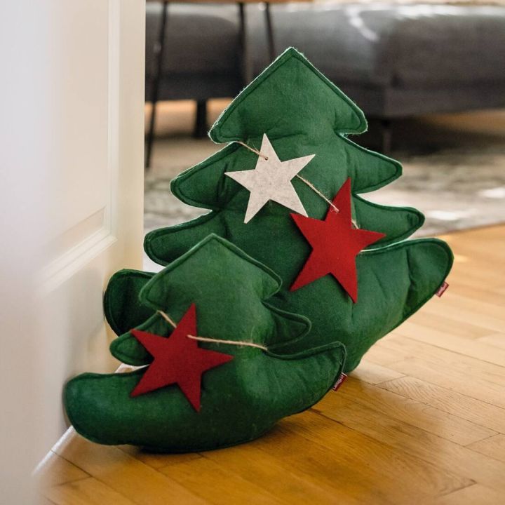 Zwei grüne, weihnachtliche Kissen in Form von Tannenbäumen mit roten und weißen Sternen dekoriert, stehen auf einem Holzboden vor einer offenen Tür in einem Wohnzimmer.