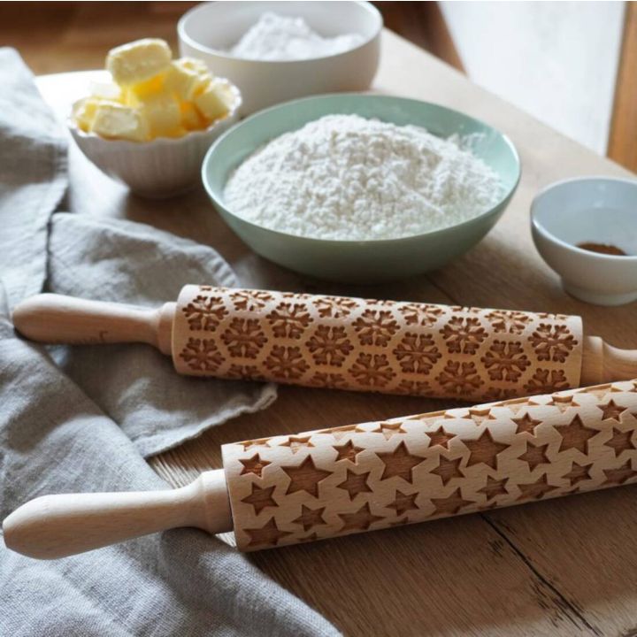 Zwei Holz-Nudelholzer mit eingravierten Schneeflocken- und Sternmotiven, umgeben von Zutaten wie Butter, Mehl und Zimt, auf einem Holztisch.