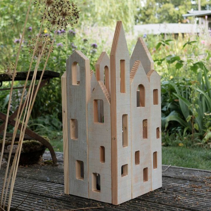 Holzlaterne in Form eines mehrstöckigen Hauses mit ausgeschnittenen Fenstern auf Terrasse, umgeben von Pflanzen im Garten.