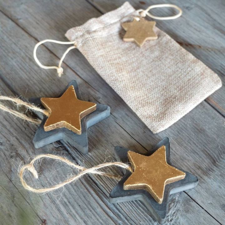 Zwei graue Sterne mit goldenen Sternen darauf liegen auf einer Holzoberfläche. Ein brauner Stoffbeutel mit einem Kordelzug und einem kleinen goldenen Sternanhänger liegt daneben.