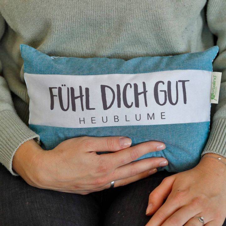 Person hält blaues Kissen mit weißem Band, darauf steht in schwarzen Buchstaben "Fühl Dich Gut – Heublume". Kissen hat kleines Etikett an der Seite. Hände umschließen das Kissen.