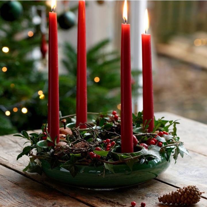 Ein Adventskranz mit vier brennenden roten Kerzen, dekoriert mit grünen Zweigen, roten Beeren und Tannenzapfen, steht auf einem Holztisch. Im Hintergrund ist ein Weihnachtsbaum zu sehen.