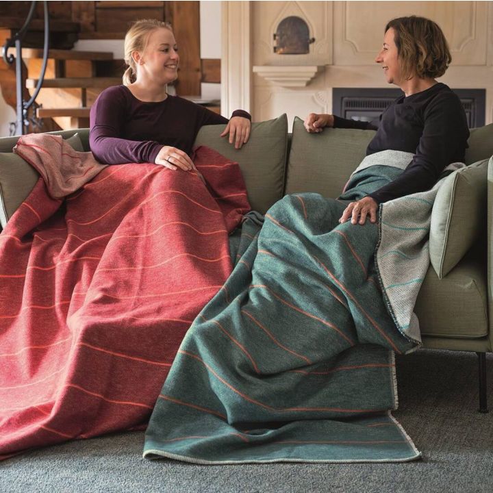 Zwei Personen sitzen l&auml;chelnd auf einem Sofa, jeweils in rote und gr&uuml;ne Decken geh&uuml;llt. Der Raum ist elegant eingerichtet mit Kamin im Hintergrund.