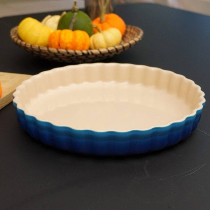 Runde Tarteform aus Keramik mit gewelltem Rand, innen cremefarben und außen in Blautönen verlaufend. Die Form steht auf einer dunklen, glatten Oberfläche. Im Hintergrund befindet sich ein geflochtener Korb mit verschiedenfarbigen Zierkürbissen in Gelb, Or