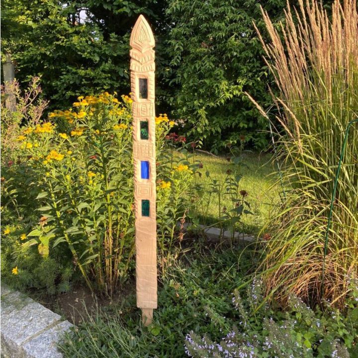 Holzskulptur mit geometrischen Mustern und farbigen Glasfenstern in einem Gartenbeet, umgeben von gelben Blumen und hohen Gräsern, im Hintergrund grüne Büsche.