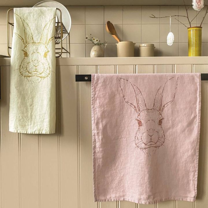 Zwei Geschirrtücher mit Hasenkopf-Aufdruck hängen in einer Küche; eines in Hellgrün mit gelbem Motiv, das andere in Rosa mit braunem Motiv, vor beigefarbener Wandverkleidung mit Küchenutensilien im Hintergrund.