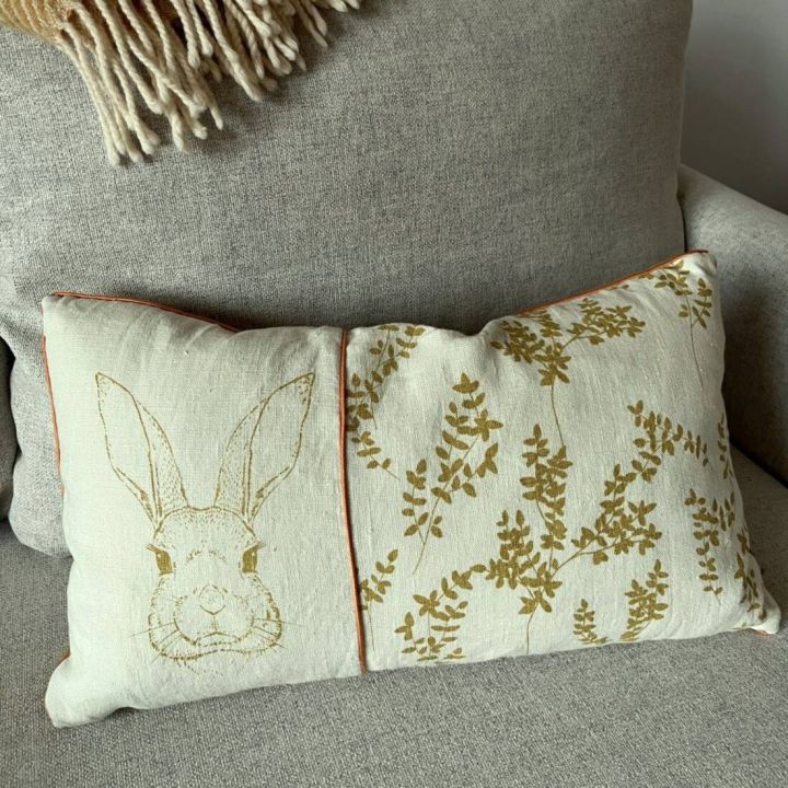 Rechteckiges Zierkissen auf grauem Sofa, mit naturfarbenem Bezug. Links ist eine skizzierte Hasenzeichnung, rechts ein Muster aus goldfarbenen Bl&auml;ttern. Oben liegt eine beigefarbene, gefranste Decke.