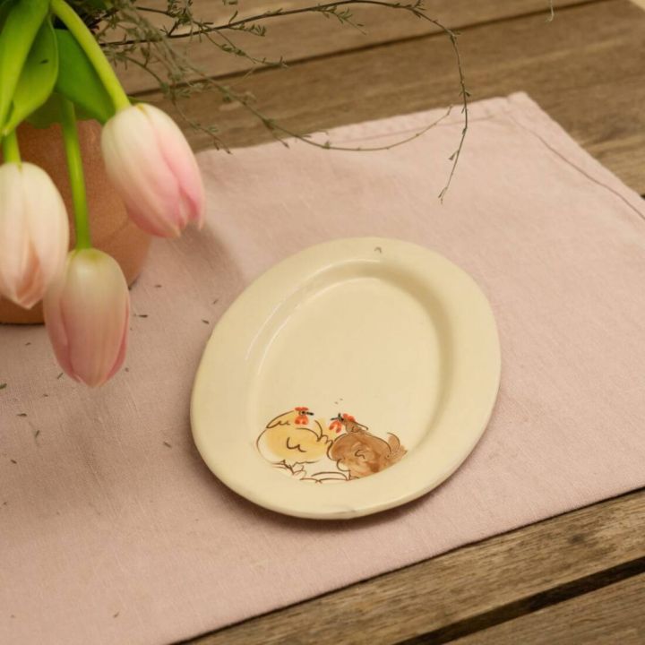 Beige ovale Keramikschale mit Hühnerillustration, auf einem hellrosa Tischläufer neben Tulpen in Vase.