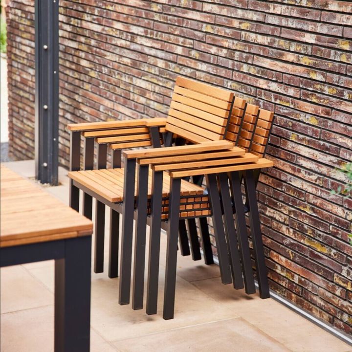 Im Bild sind mehrere stapelbare Stühle aus Holz und Metall zu sehen. Sie stehen nebeneinander an einer gemauerten Wand im Freien. Ein Teil eines Tisches aus denselben Materialien ist im Vordergrund sichtbar.