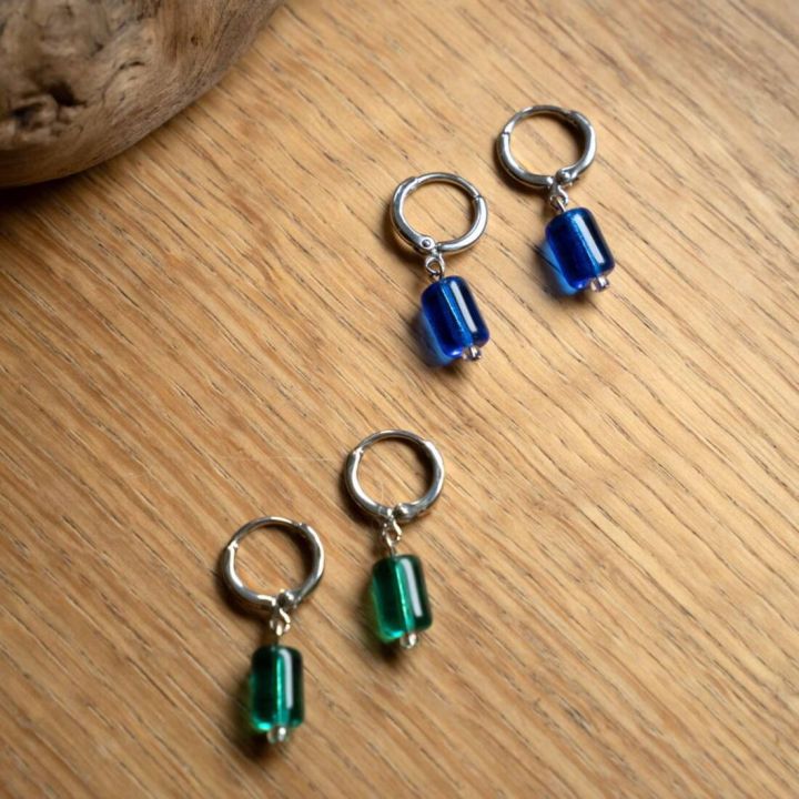 Silberne Ohrringe mit runden Fassungen und hängenden, zylindrischen Glasanhängern in Blau und Grün, auf hellem Holzhintergrund platziert.